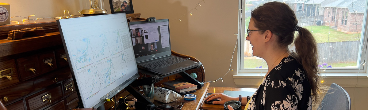 Woman working at a desk in a home office with a large computer monitor and laptop open beside it. The woman is smiling; the monitor is showing weather model displays; the laptop is showing a virtual meeting.