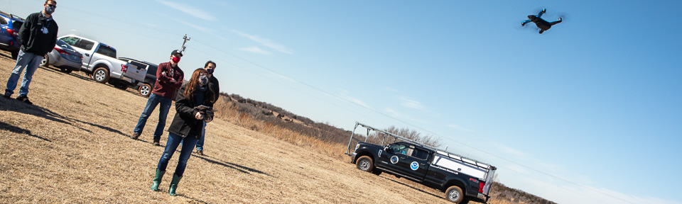 Four people flying a drone over a field. Pickup truck in the background with NOAA logos on it
