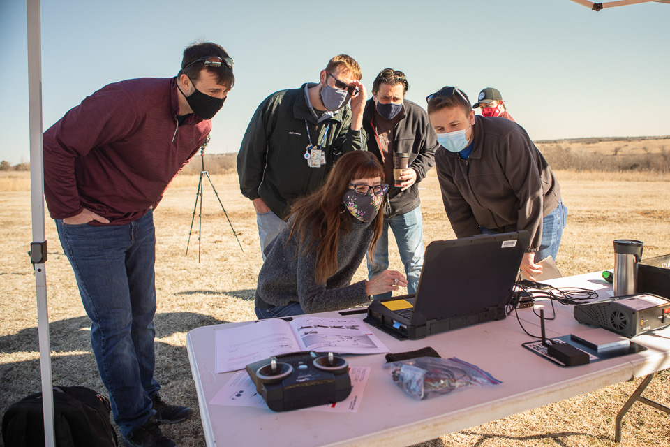 Six people wearing masks, gathered around a laptop beneath a canopy on a sunny day