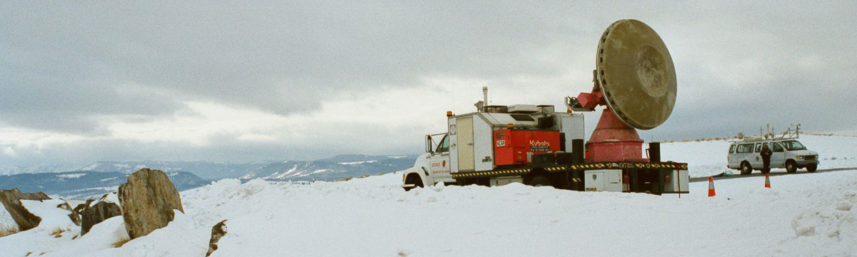 Radar mounted on flatbed truck, parked beside a road in the snow. Mountains are visible in the background. An instrumented van is parked nearby with a person standing by it.