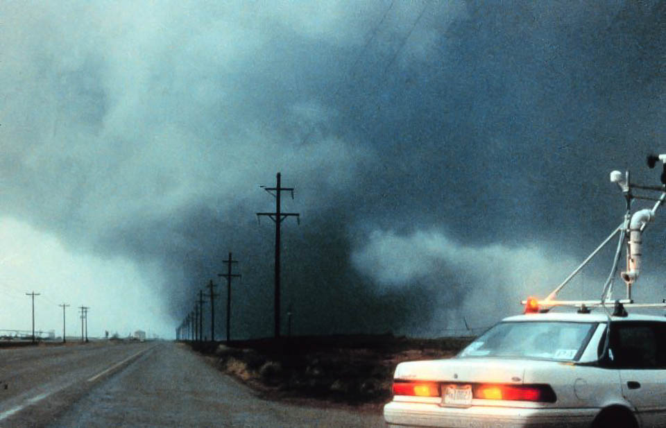 90s-era sedan with weather observing equipment mounted to the roof, stopped by the side of a rural highway. A large tornado is in the background.