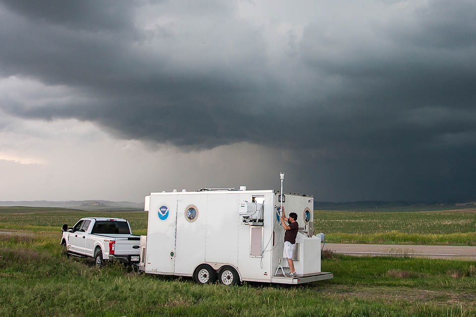 White utility trailer attached to white pickup truck in a grassy area near a rural highway. The sky is dark and cloudy. A man is working on equipment mounted to the rear of the trailer.