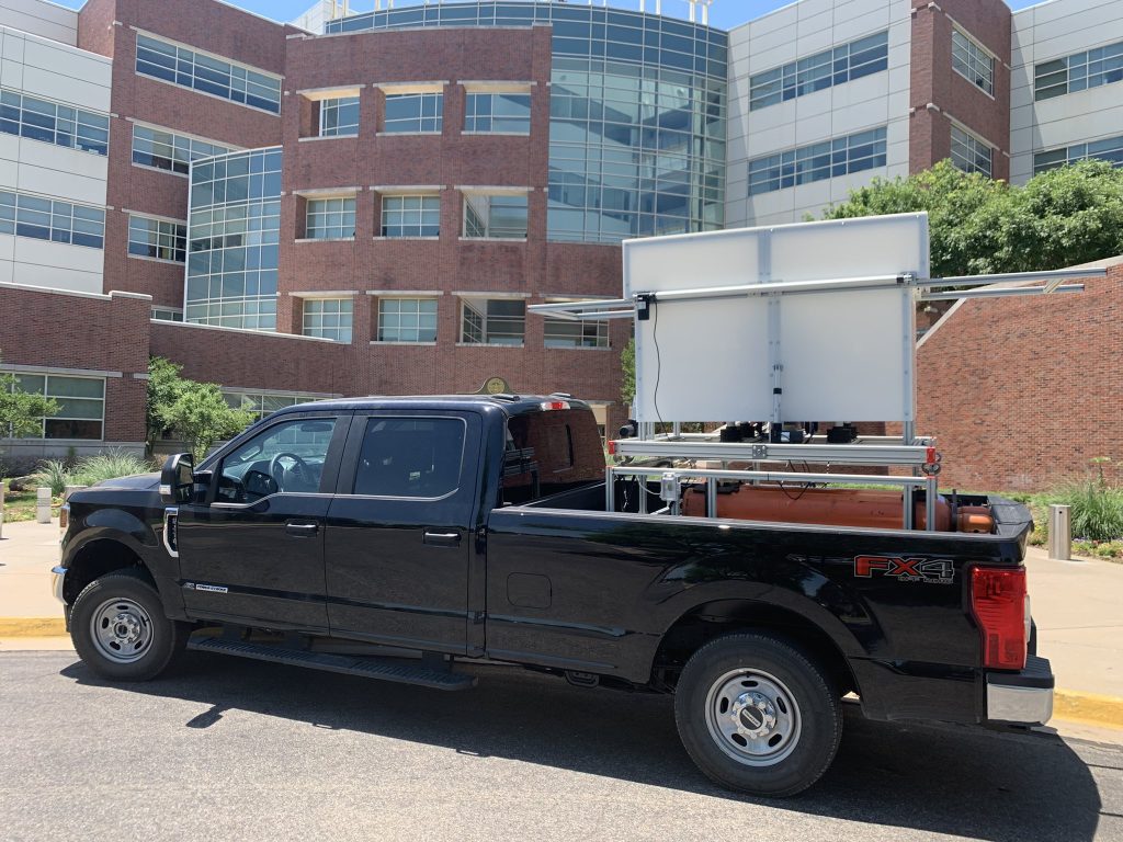 Black pickup truck with white electronics container in the bed. The truck is parked in front of a large brick office building