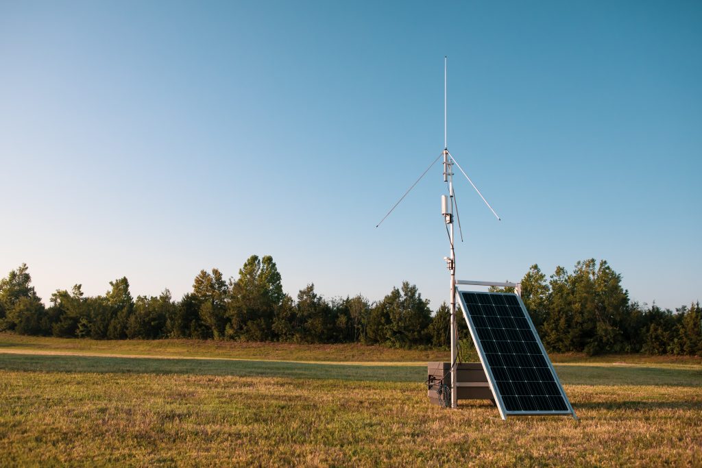 Metal pole holding electronic equipment and an antenna. Beside it is a solar panel. They are standing in an open grassy area with trees visible in the background